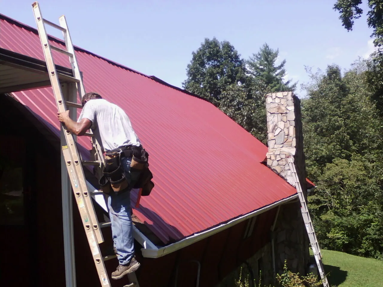 Roofer on a ladder beside a red metal roof and stone chimney — Soffit & Fascia Repair by Roofing Brothers in Oakland